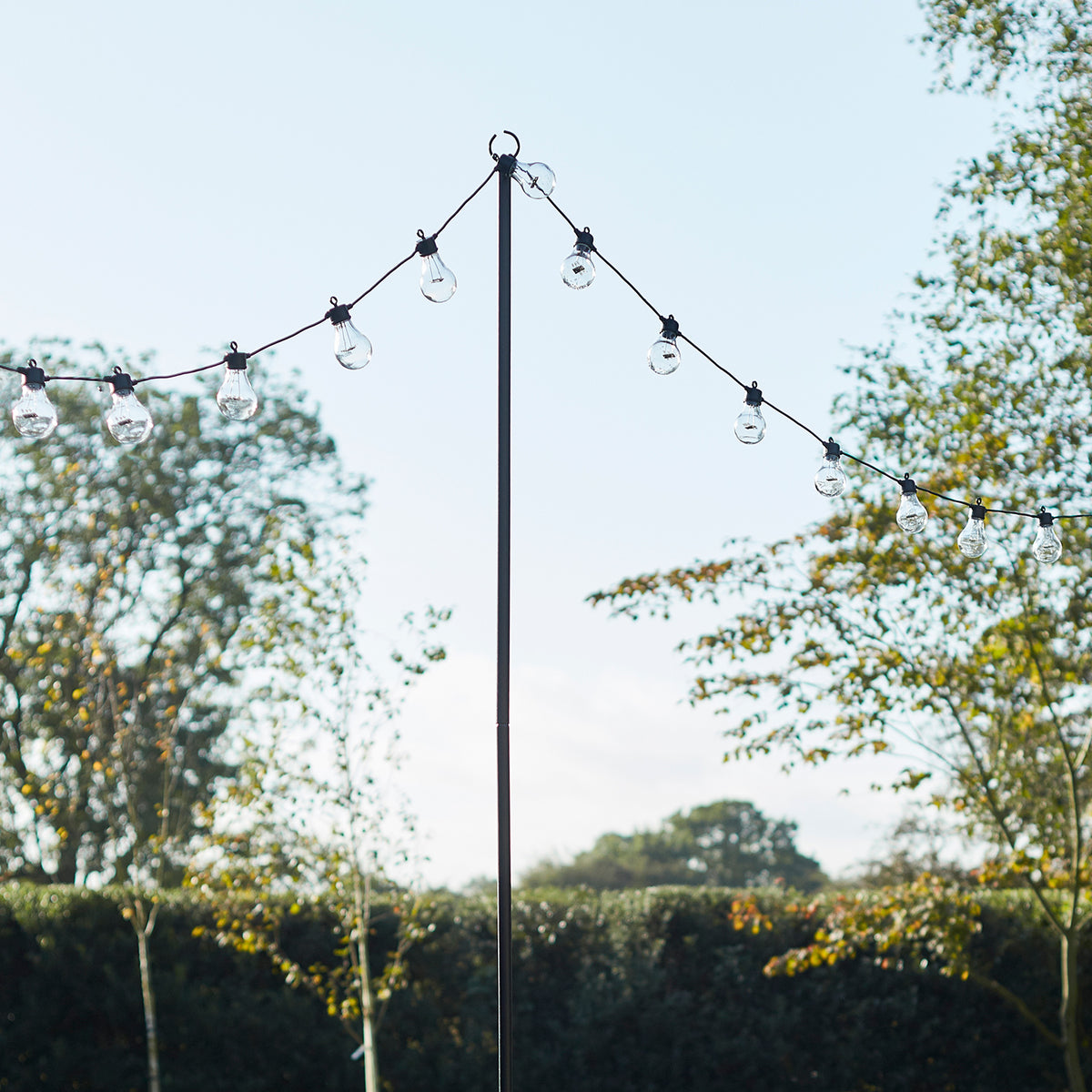 Schwarze Lichterkettenstange mit aufgehängter Glühbirnen-Lichterkette im Garten vor blauem Himmel