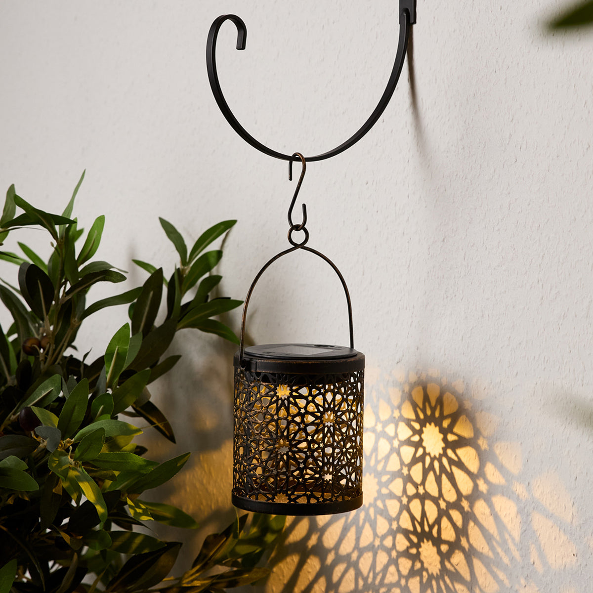 Schwarze Laterne mit Musterlicht hängt an Metallhaken an weißer Wand mit Pflanzen im Hintergrund