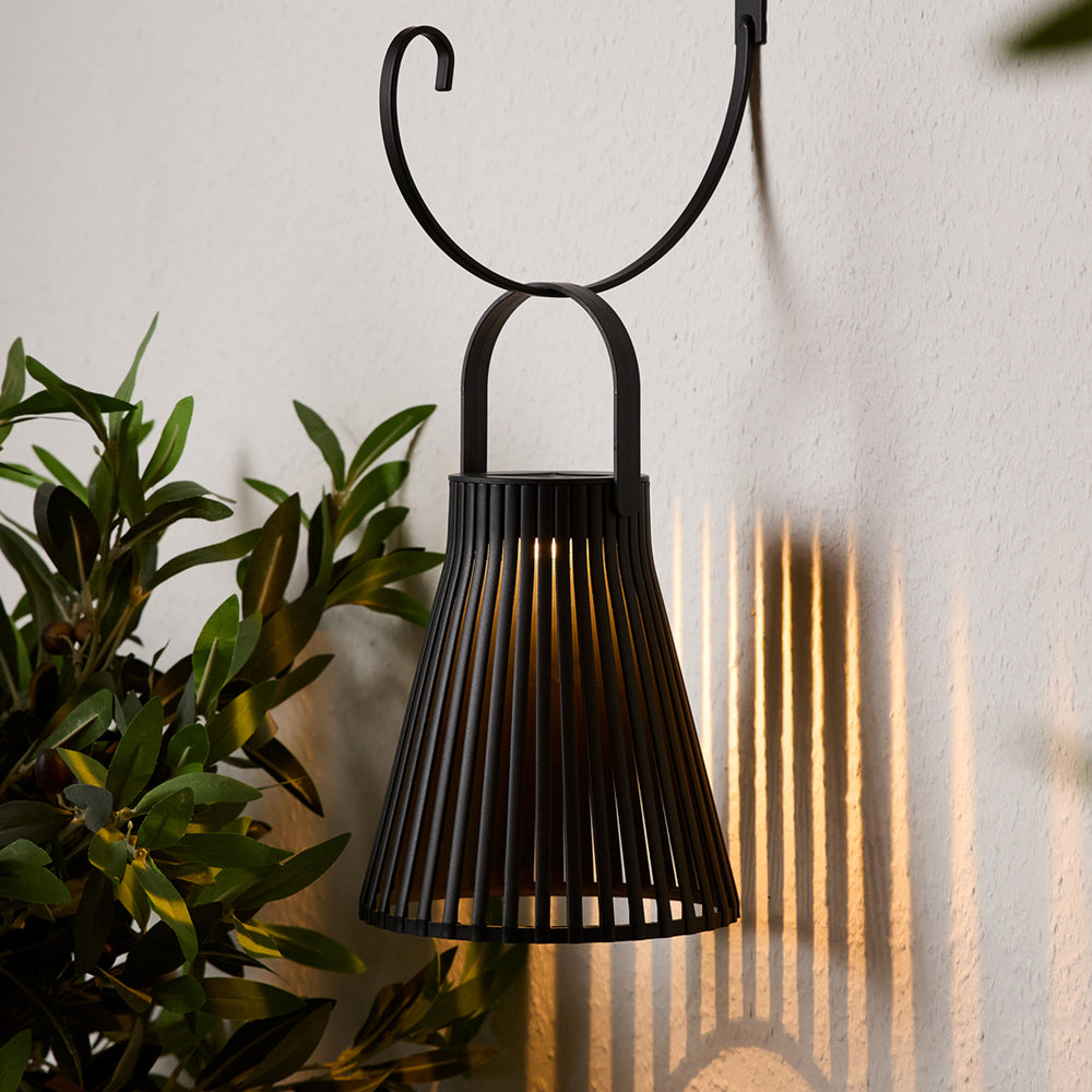 Schwarze Laterne hängt an schwarzem Metallhaken an weißer Wand mit Pflanzen im Hintergrund