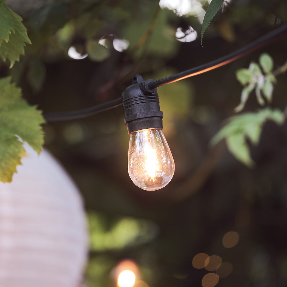 Nahaufnahme einer warmweiß leuchtenden Glühbirne an schwarzem Kabel im Garten