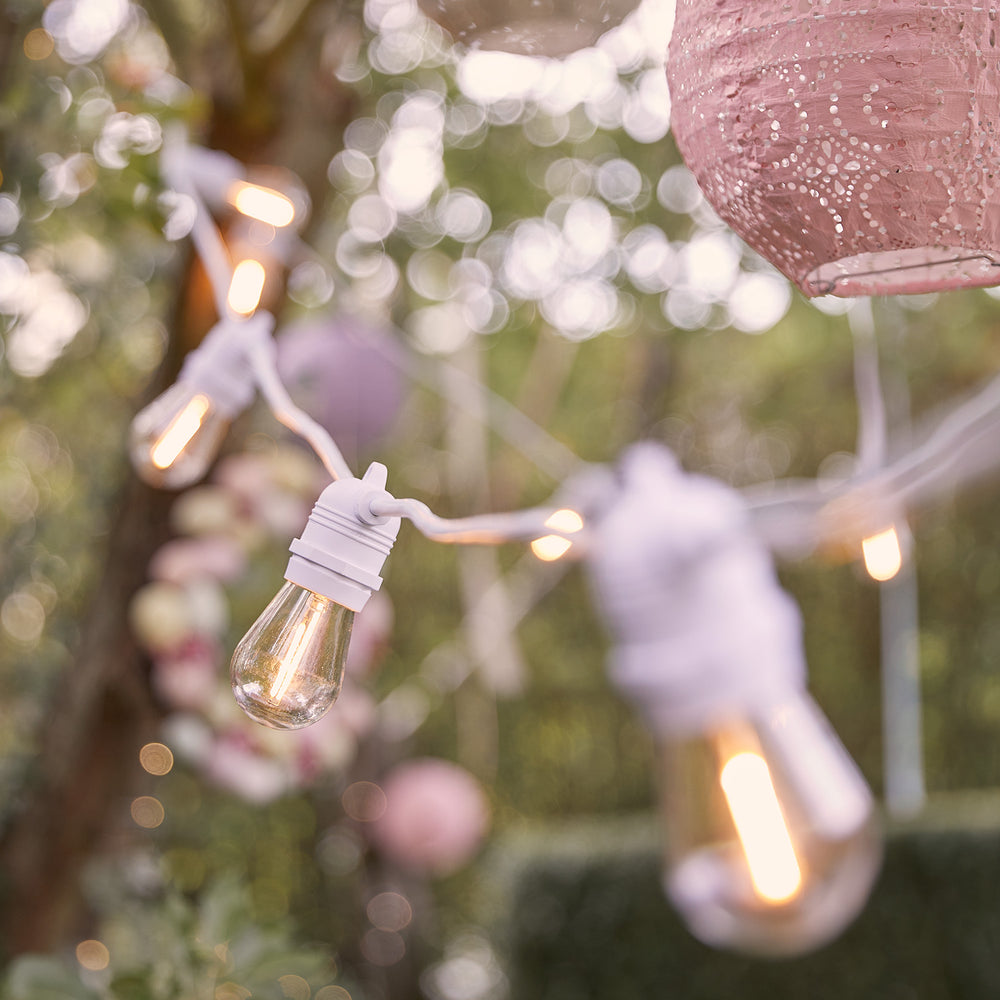 Lichterkette mit warmweißen Glühbirnen an weißem Kabel, dekoriert mit rosa Lampions im Garten