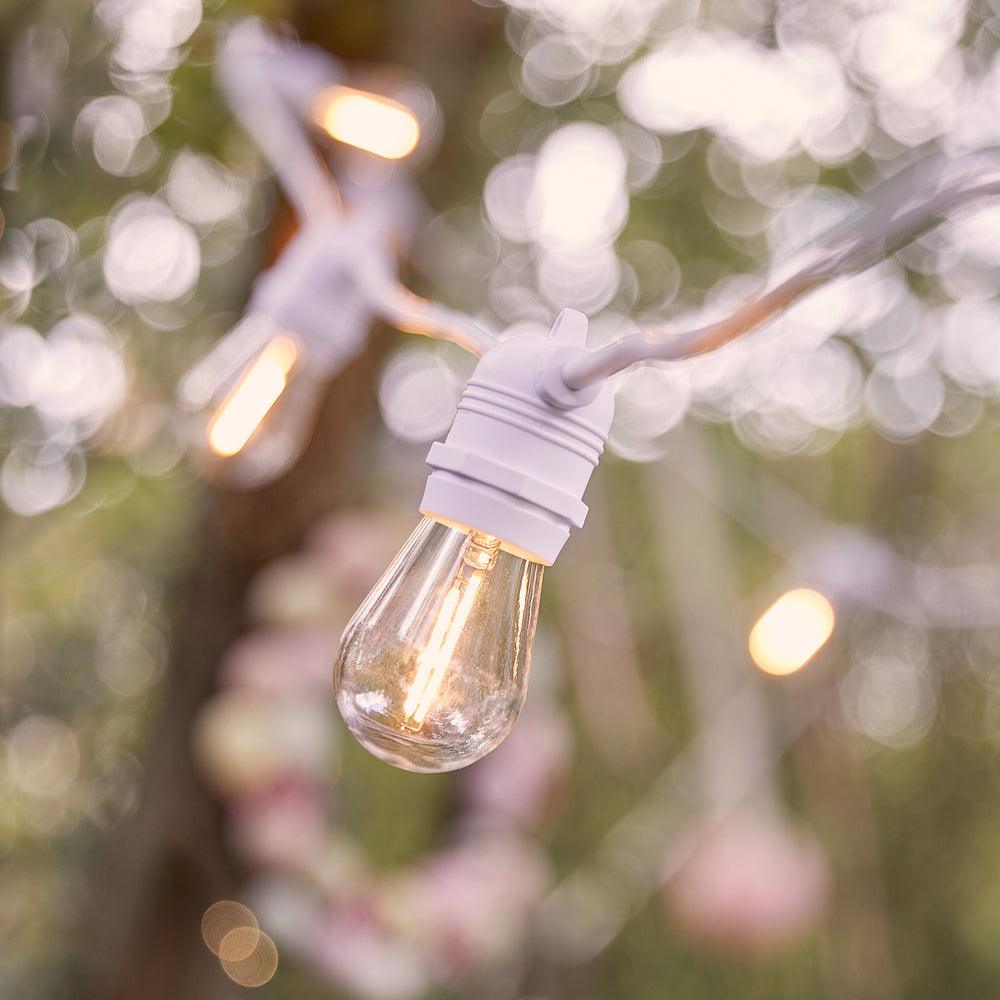 Nahaufnahme einer warmweißen LED-Glühbirne an weißem Kabel vor unscharfem Gartenhintergrund