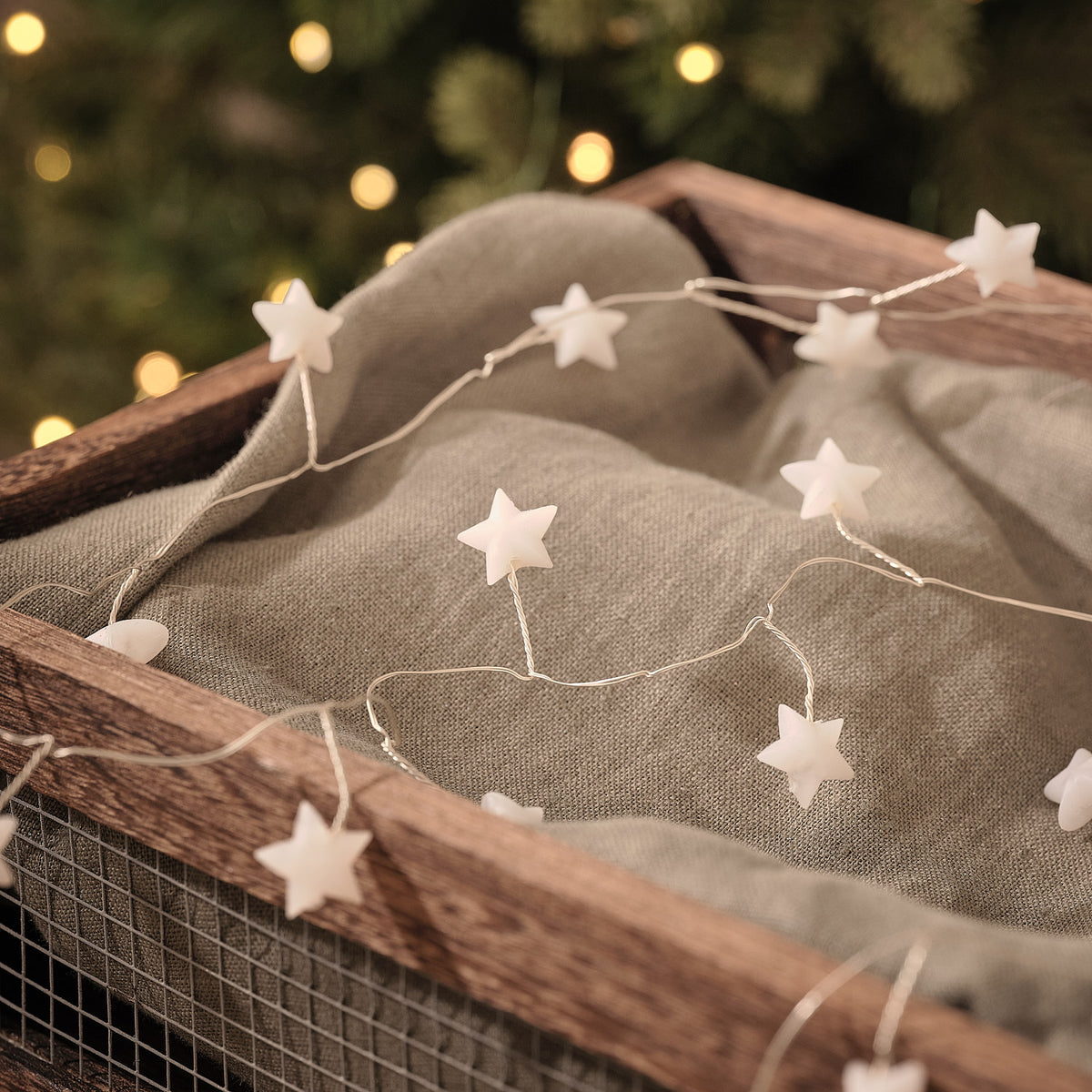 Weiße Stern-LED-Lichterkette mit mehreren leuchtenden Sternen auf beigefarbenem Stoff und Holzast, warmweiße LED-Lichter, bokeh Hintergrund mit Weihnachtsbaum.