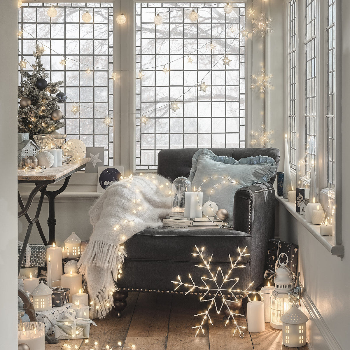 Fenster-Weihnachtsdekoration mit weißer Stern-LED-Lichterkette, Weihnachtsbaum und Schneeflocken-Lichtern, Wohnzimmer mit grauem Sofa, warmweiße Weihnachtsbeleuchtung am großflächigen Fenster.