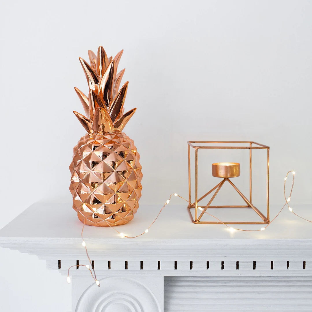 Kupferfarbene Ananasfigur und geometrischer Kerzenhalter mit LED-Lichterkette auf weißem Kaminsims