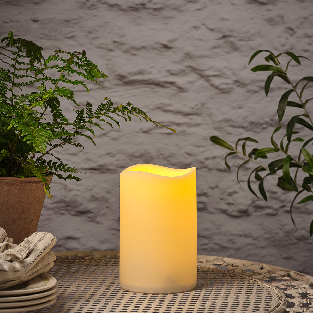 Leuchtende LED Outdoor Kerze auf Gartentisch mit Pflanzen im Hintergrund