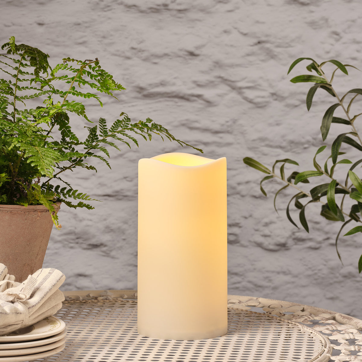 LED Outdoor Kerze mit warmem Licht auf Tisch neben Pflanzen und Geschirr