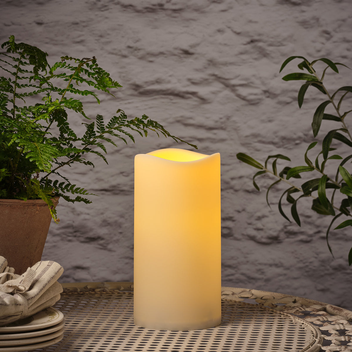 Leuchtende LED Outdoor Kerze auf Metalltisch mit Pflanzen im Hintergrund