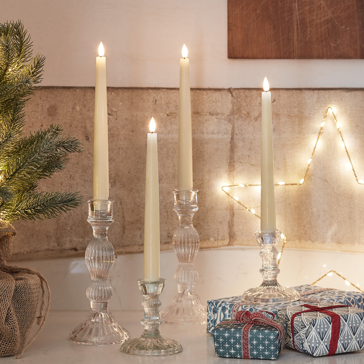 Drei LED Stabkerzen in transparenten Glas-Kerzenhaltern vor einer Wand mit Stern-Lichterkette und Geschenken