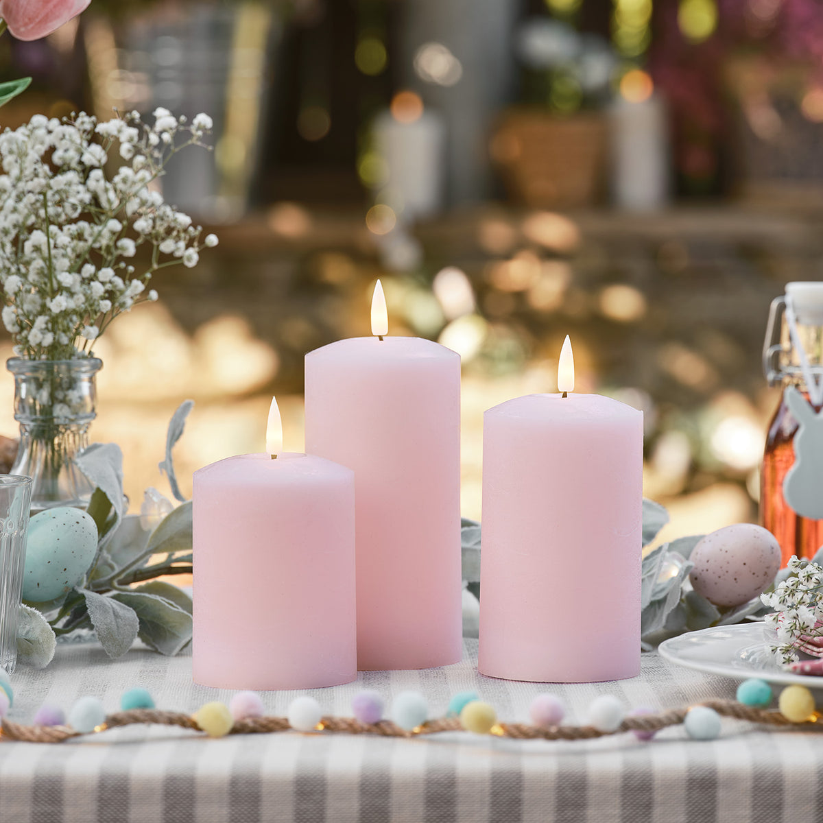 Drei rosa LED Stumpenkerzen mit Osterdeko auf gedecktem Gartentisch