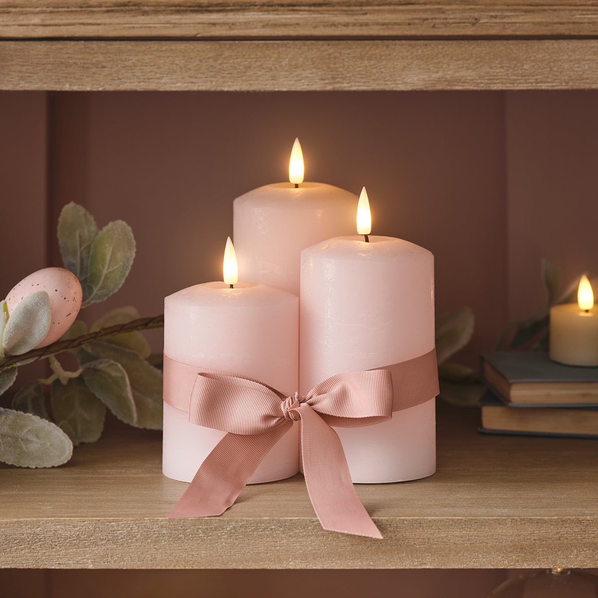 Drei rosa LED Stumpenkerzen mit Schleife auf Holzregal mit Osterdeko