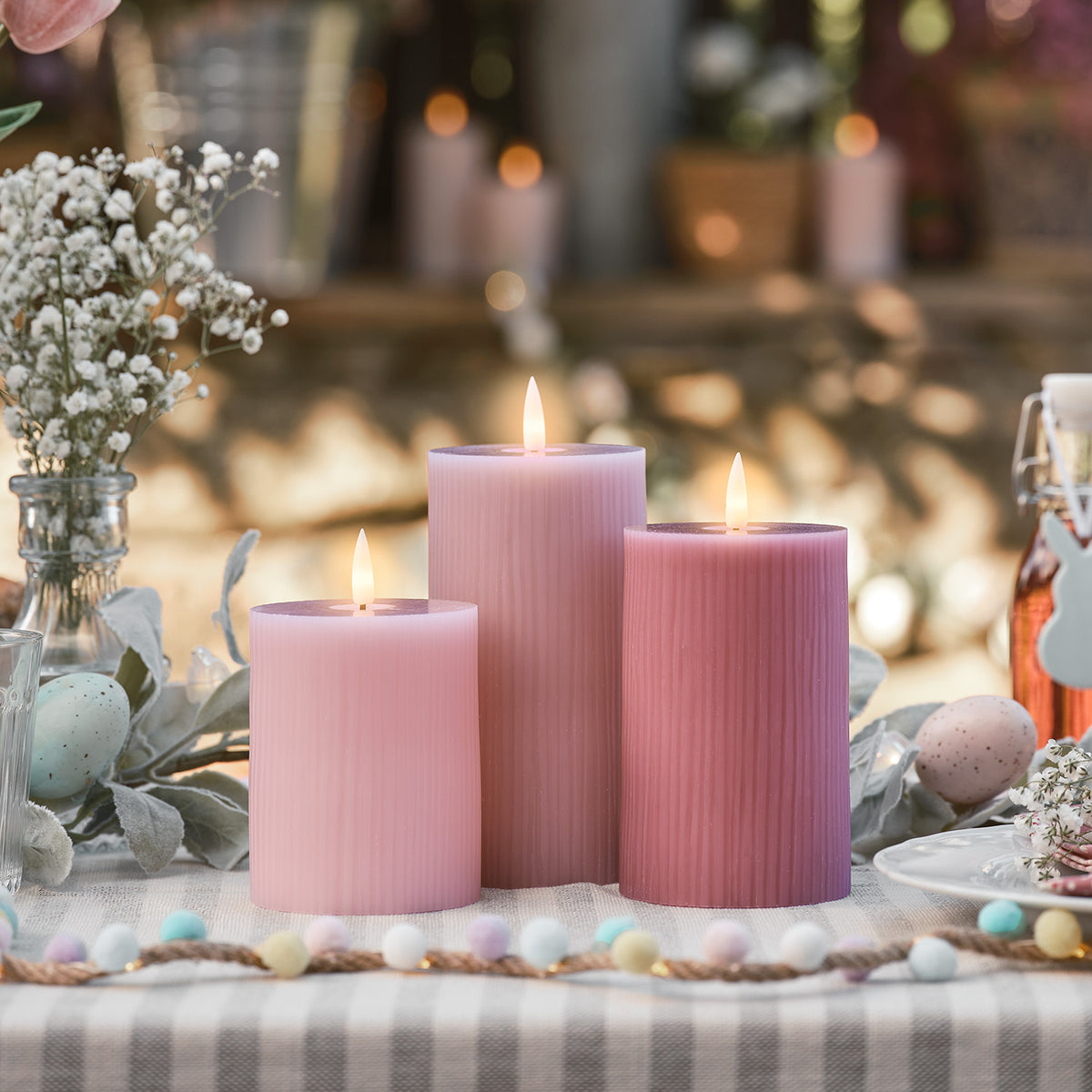 Drei brennende LED-Kerzen in Rosa- und Pastelltönen auf gedecktem Tisch mit Osterdeko