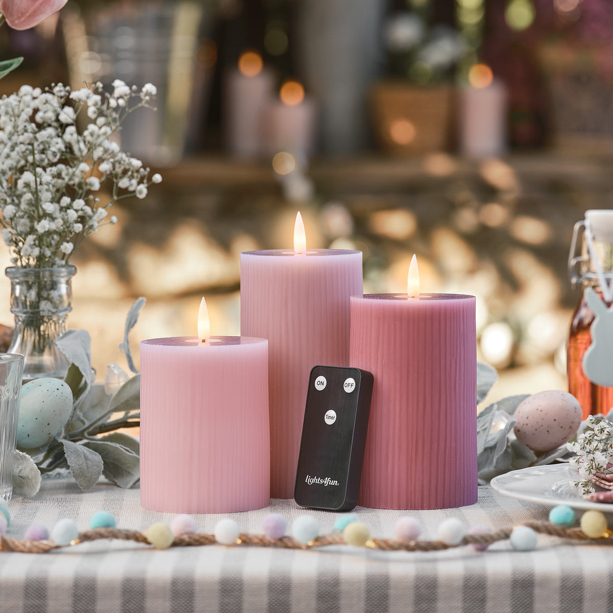 Drei brennende LED-Kerzen in Rosa- und Pastelltönen mit Fernbedienung auf Tisch mit Orangen