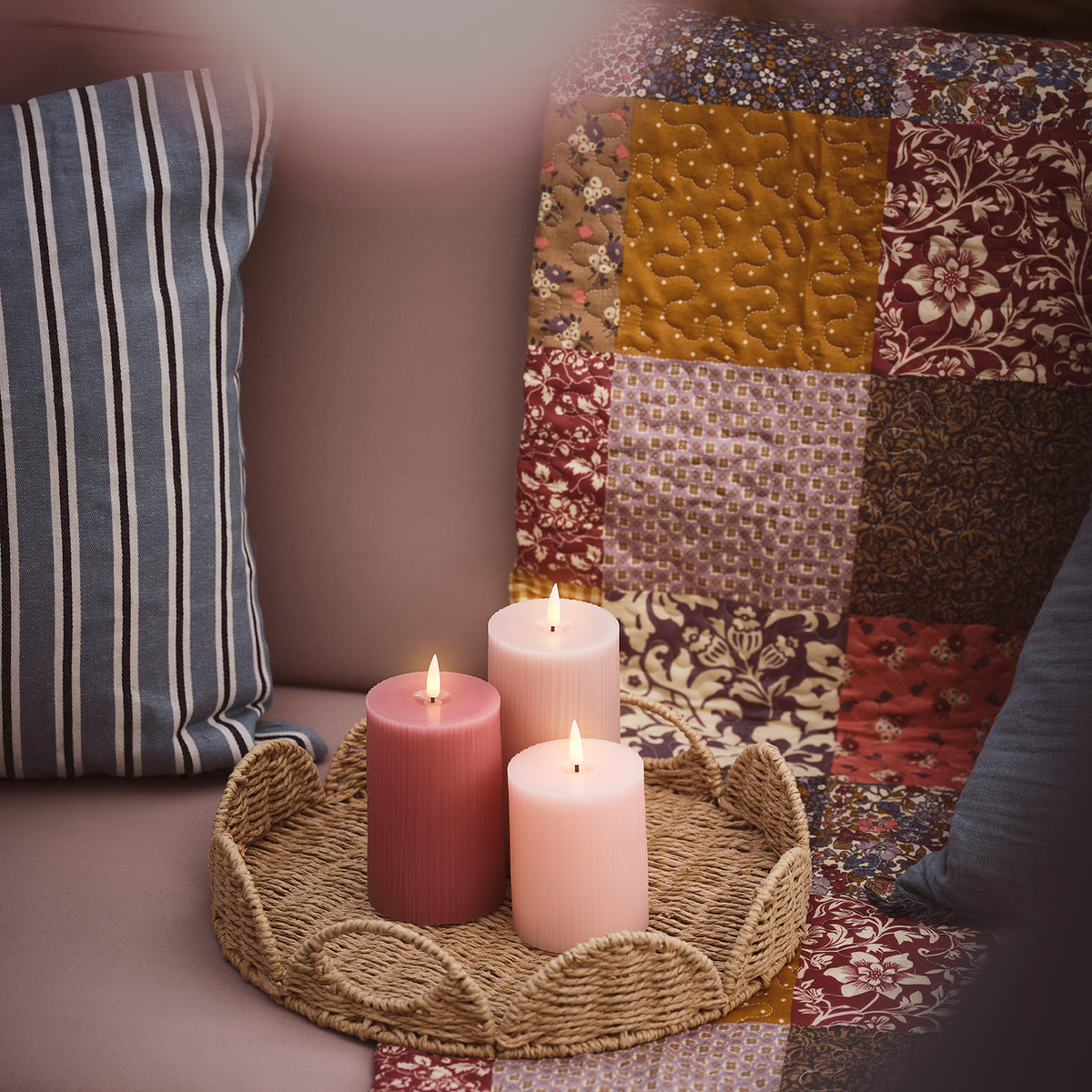 Drei brennende LED-Kerzen in Rosa- und Pastelltönen auf einem Tablett vor buntem Patchwork-Kissen