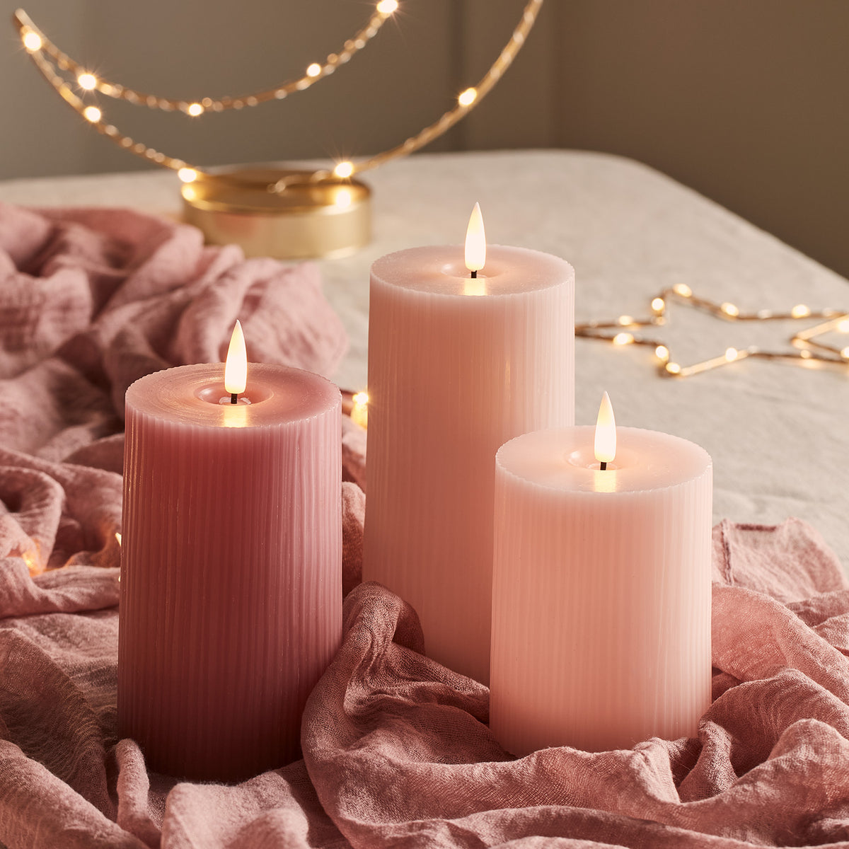 Geriffelte LED-Kerzen in Rosa- und Pastelltönen auf rosa Stoff mit Stern-Lichterkette im Hintergrund