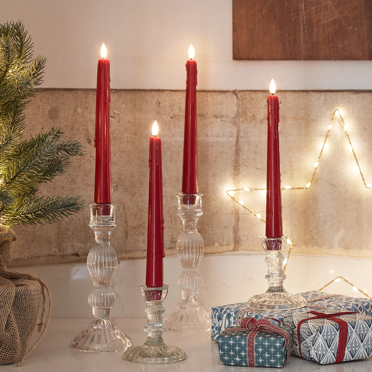 Vier rote LED Stabkerzen mit Wachstropfen in Glas-Kerzenhaltern vor Steinwand und Weihnachtsdeko