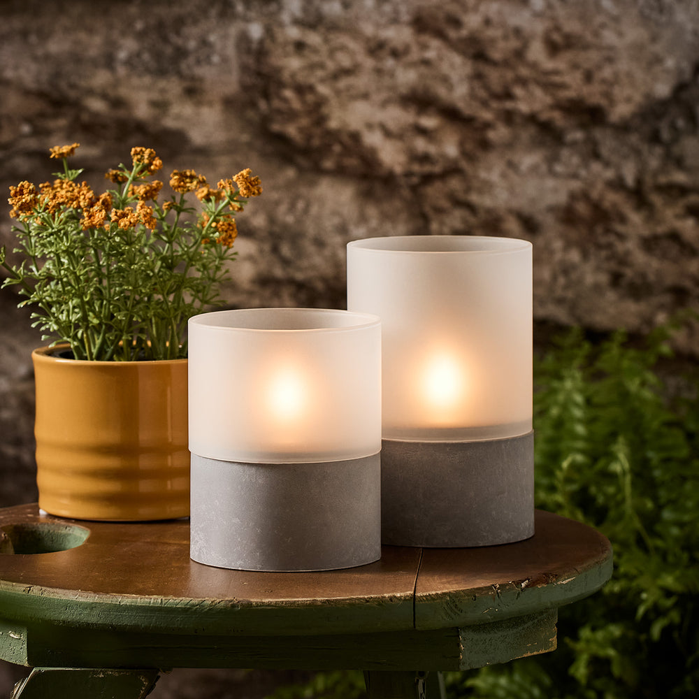 Zwei TruGlow Solar Windlichter aus Glas mit Betonfuß auf grünem Holztisch neben gelbem Blumentopf