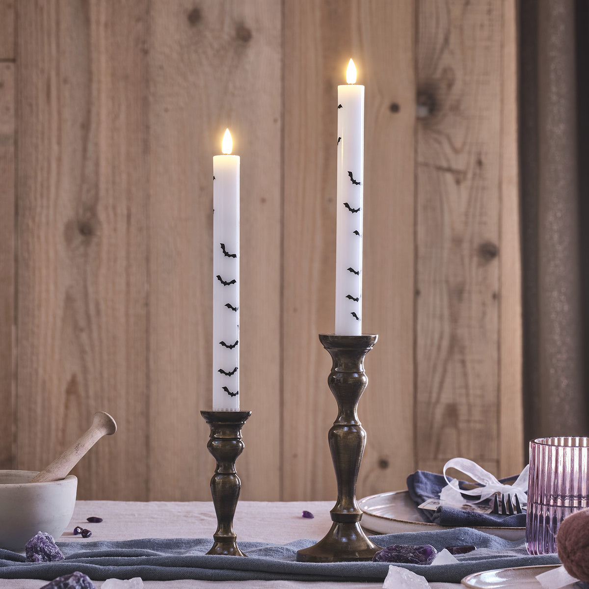 Zwei weiße LED Stabkerzen mit schwarzen Fledermausmotiven in Messingkerzenhaltern vor Holzwand