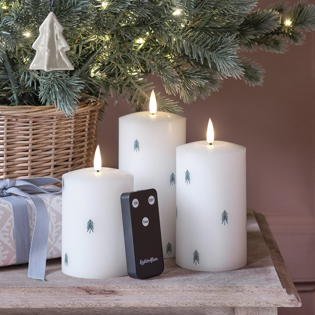 Drei weiße LED-Kerzen mit Tannenbaum-Motiv und Fernbedienung auf Holzuntergrund vor rosa Wand