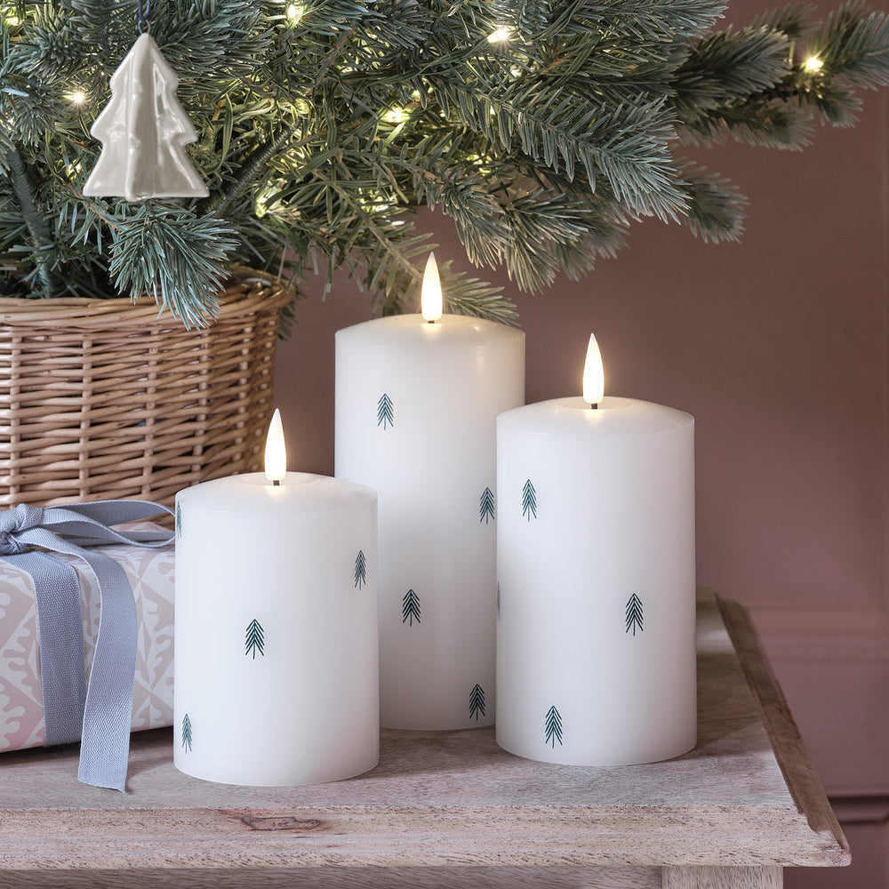 Drei weiße LED-Kerzen mit Tannenbaum-Dekor unter einem Weihnachtsbaum mit Lichterkette und Geschenk