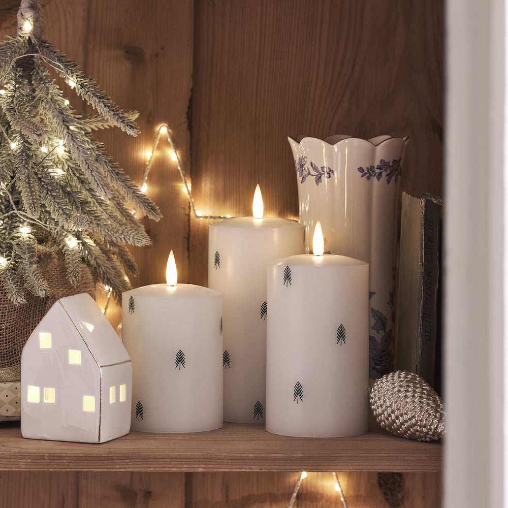 Drei weiße LED-Stumpenkerzen mit kleinen Tannenbaum-Motiven auf Holzregal neben beleuchtetem Keramikhaus