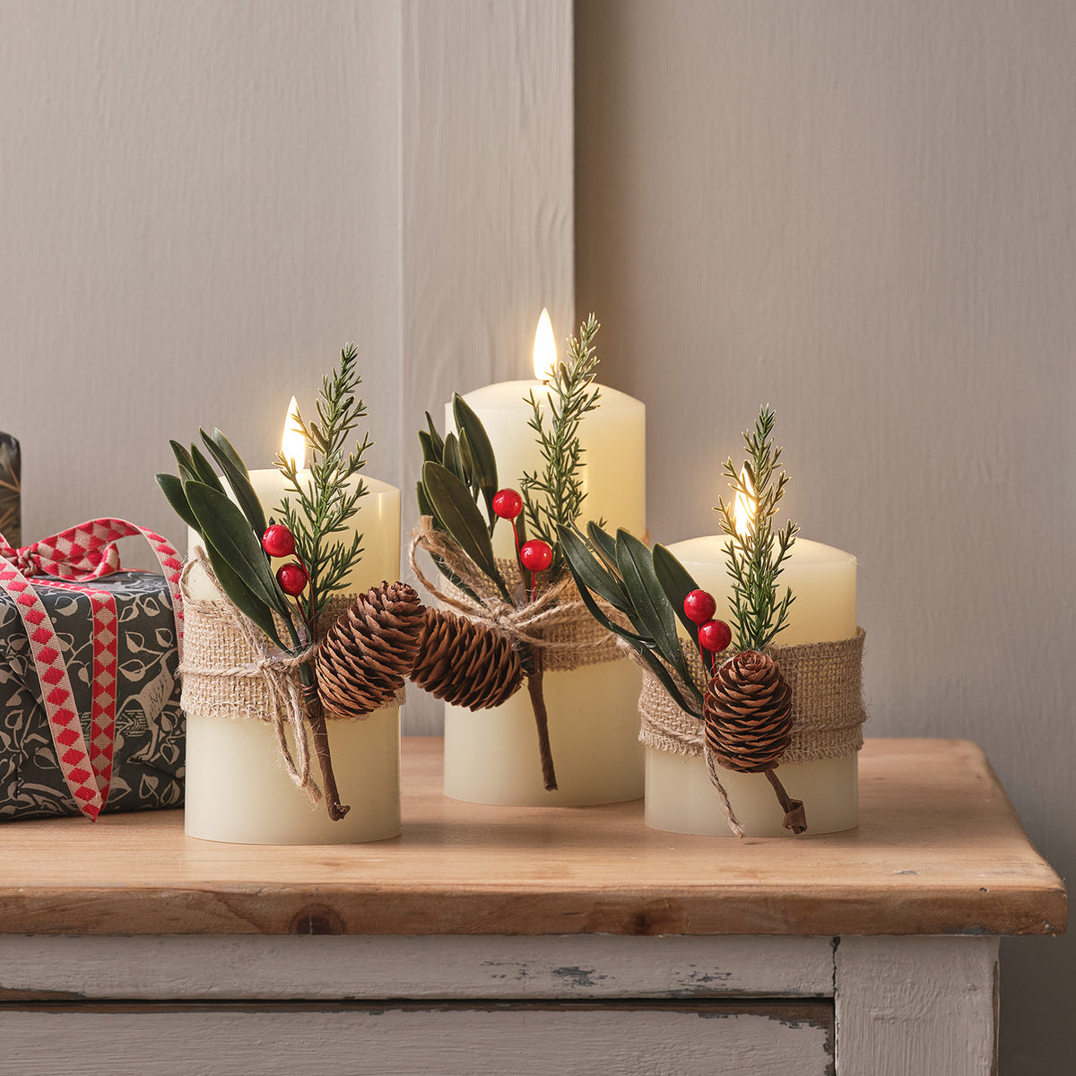 Drei cremefarbene LED Stumpenkerzen mit Winterlaub, roten Beeren und Tannenzapfen auf Holztisch