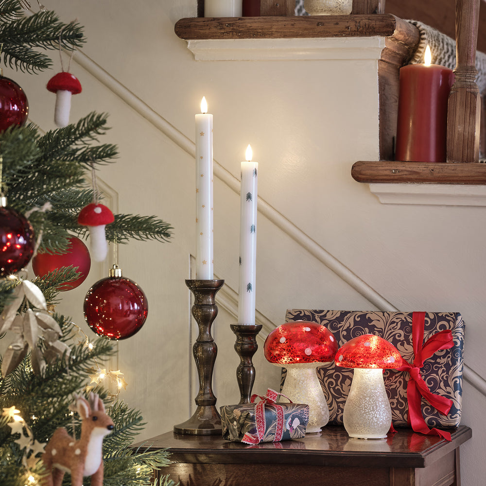 Zwei brennende LED Stabkerzen neben Weihnachtsbaum mit roten Kugeln und Pilz-Deko auf Kommode