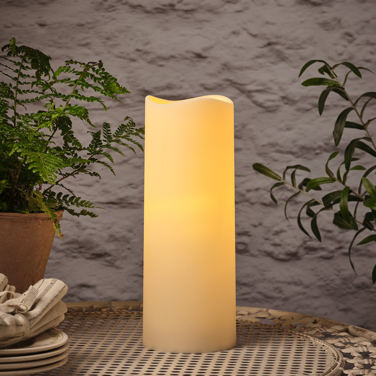 LED-Kerze für draußen mit realistischer Flamme auf Gartentisch, umgeben von grünen Pflanzen