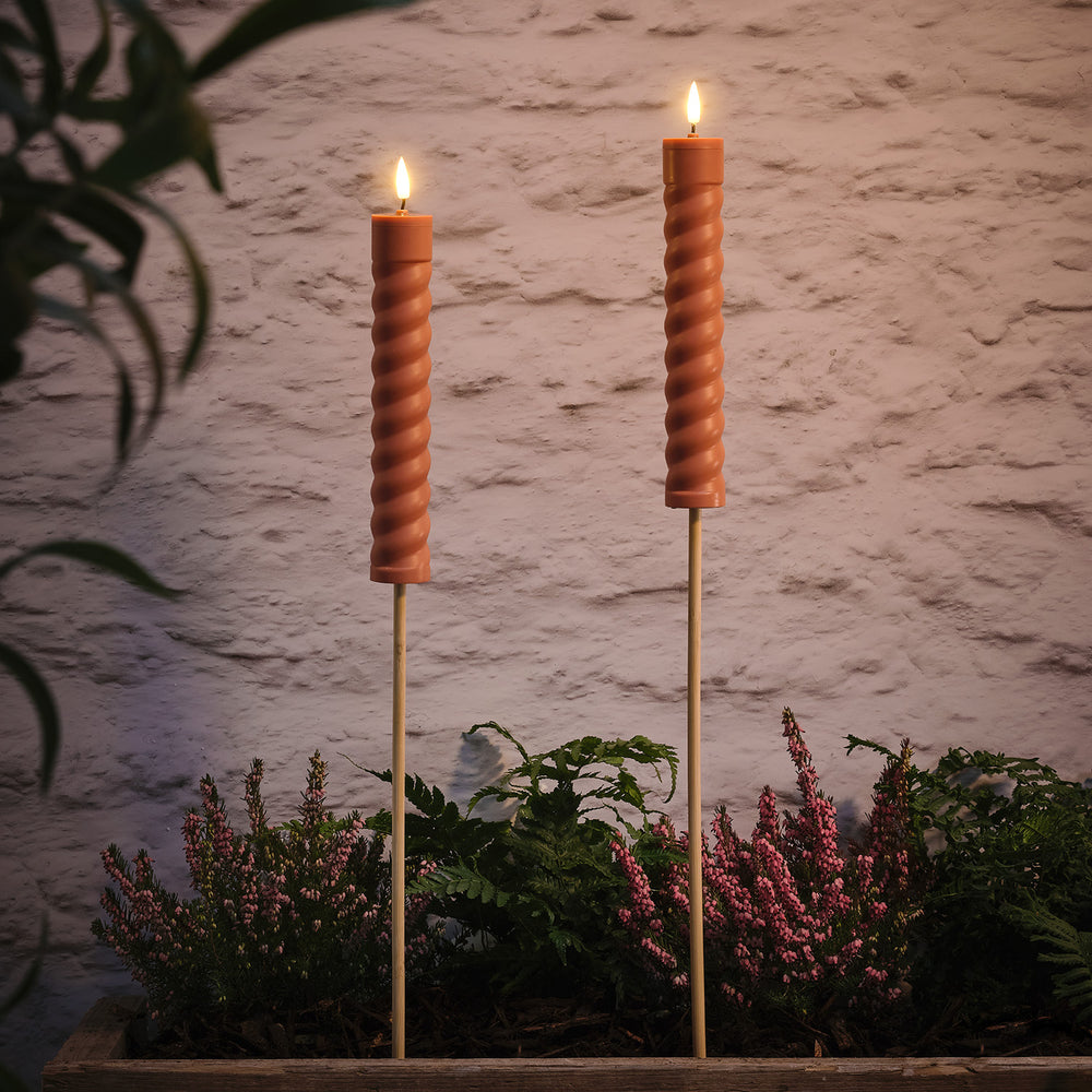 Zwei terracottafarbene Solar Stabkerzen mit LED-Flamme in einem Beet vor einer Steinwand