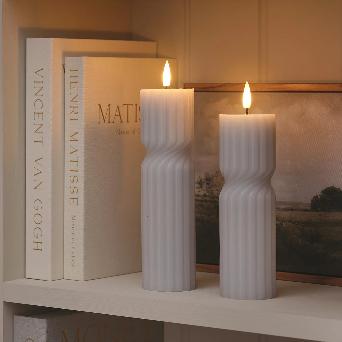 Zwei weiße LED-Stumpenkerzen mit geripptem Twist-Design auf Regal vor Büchern und Bild