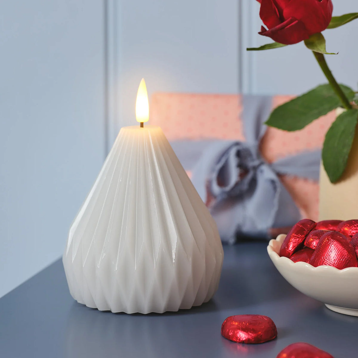 TruGlow LED-Kerze mit flackernder Flamme auf blauem Tisch neben Schale mit roten Pralinen
