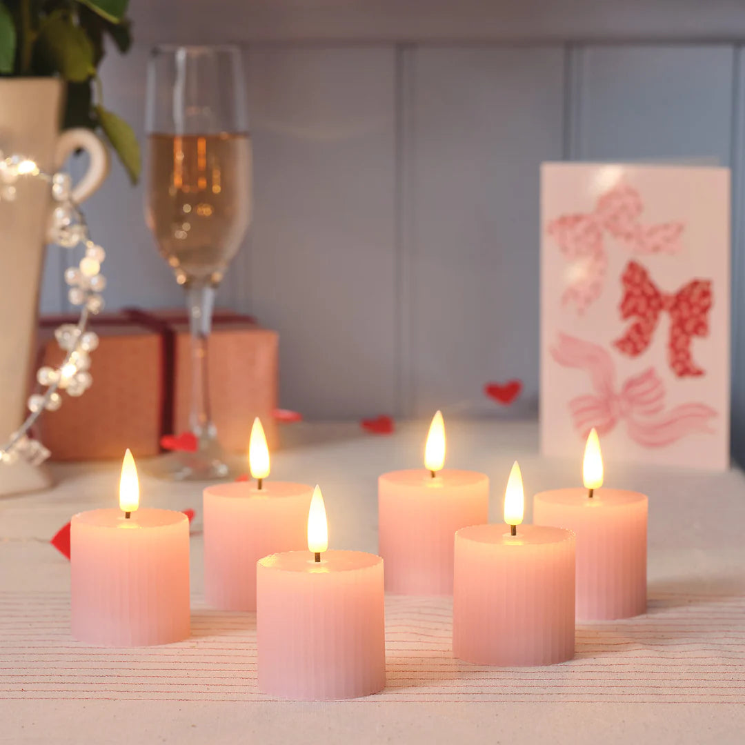 Sechs rosa LED-Votivkerzen mit flackernden Flammen auf einem Tisch mit Gläsern und Geschenken