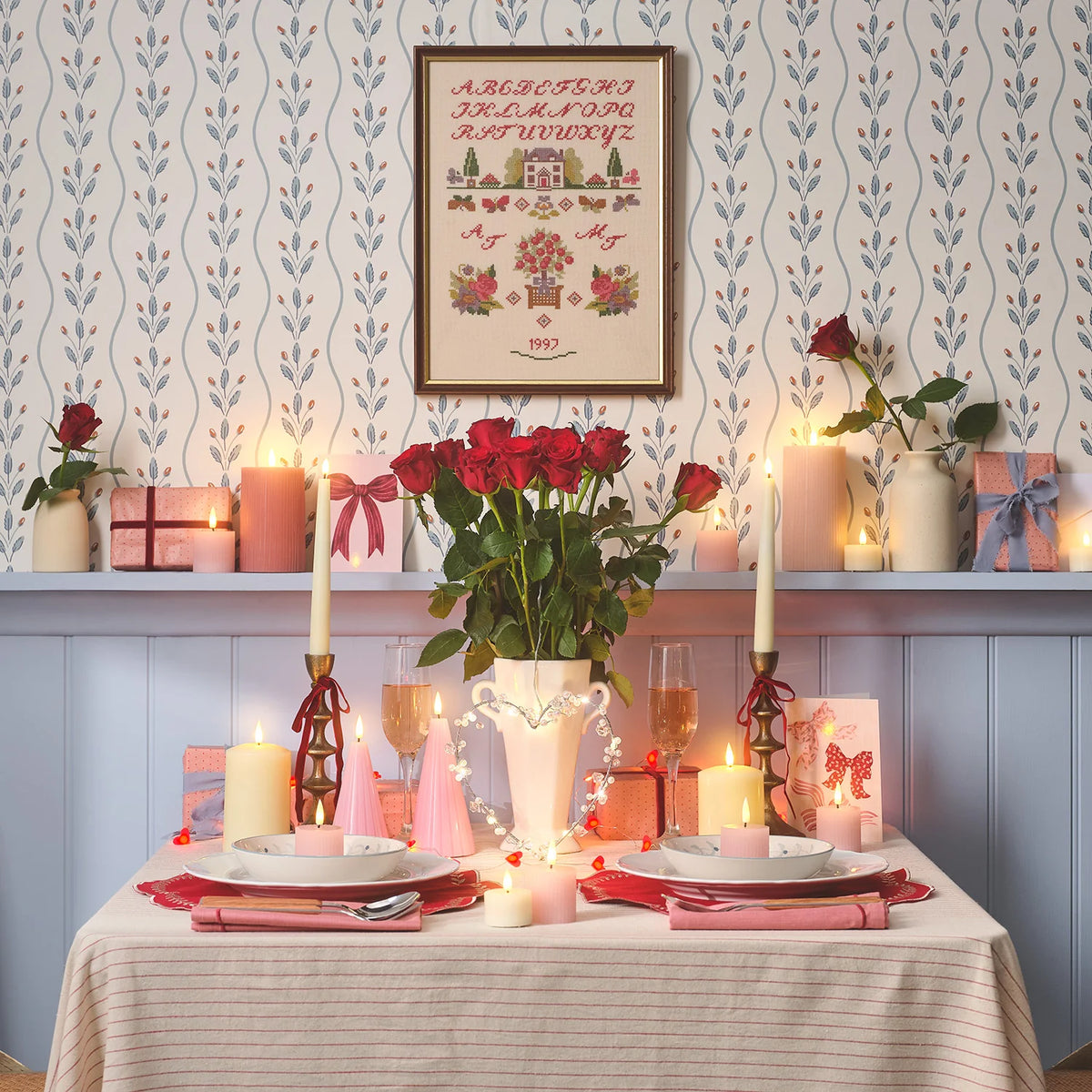 Romantisch gedeckter Tisch mit rosa LED-Kerzen, roten Rosen und Herz-Deko vor blauer Wand