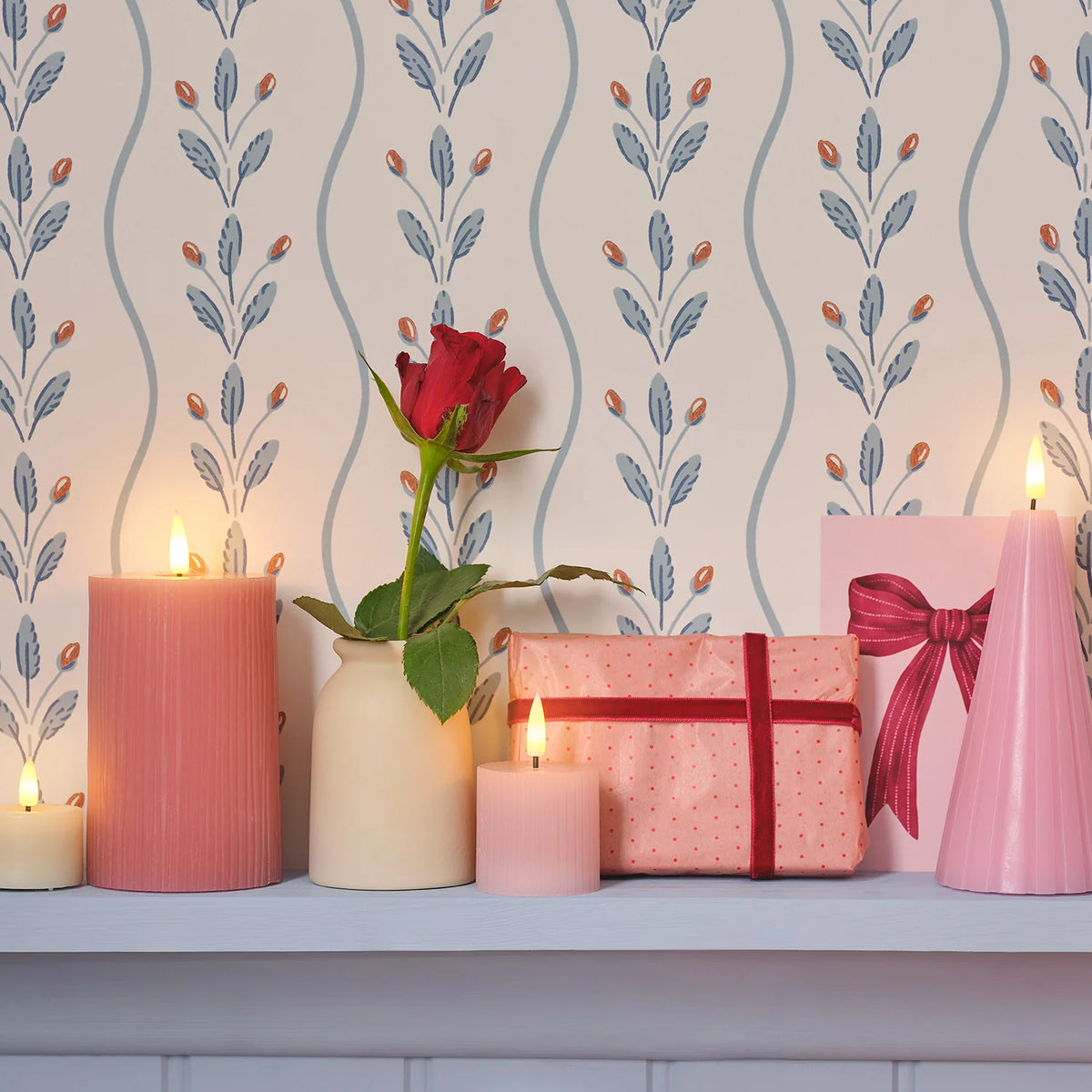 Rosa LED-Kerzen neben einer roten Rose in Vase und Geschenkpaketen vor gemusterter Tapete