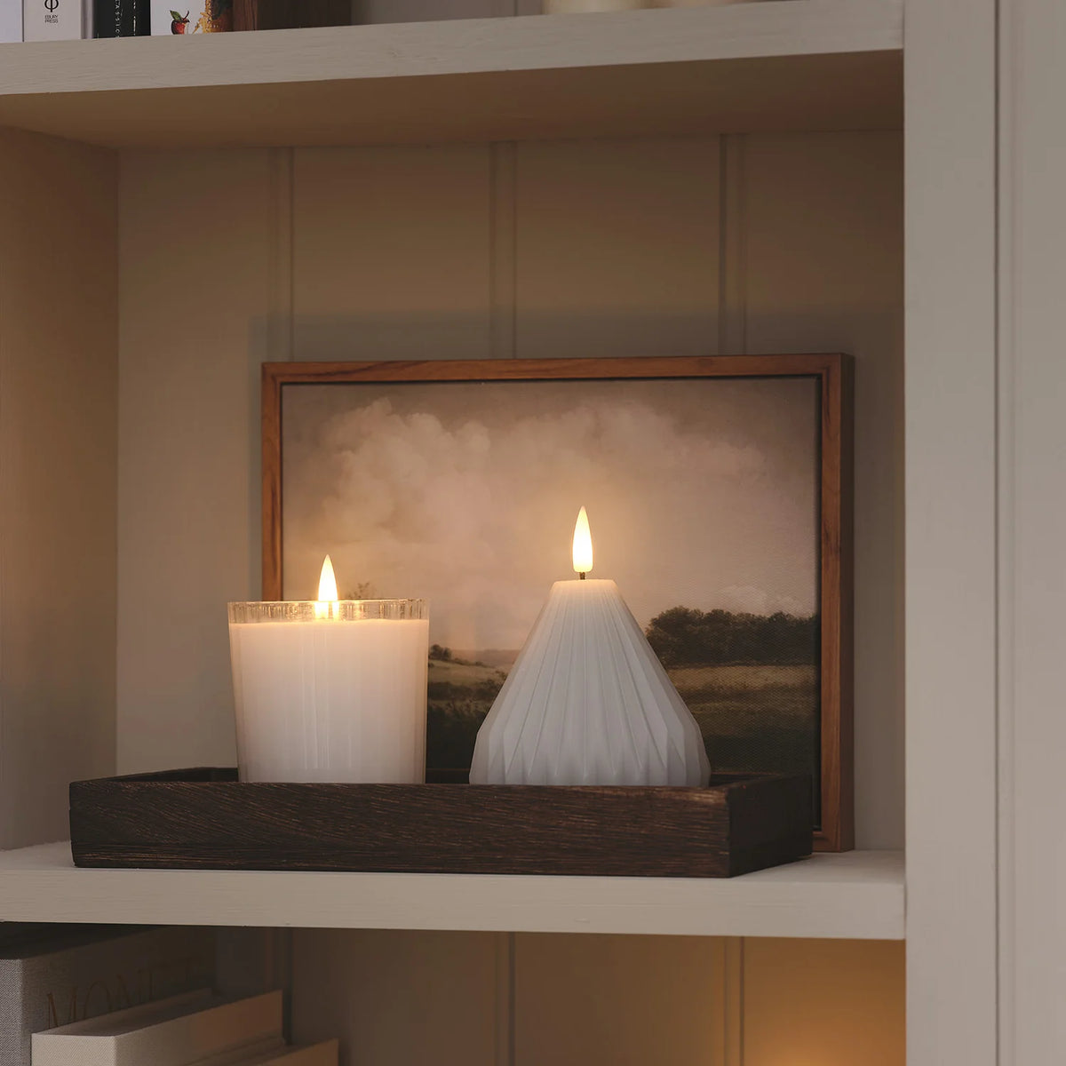 Zwei LED-Kerzen, eine im geriffelten Glas, eine kegelförmig, auf Holztablett vor Bild