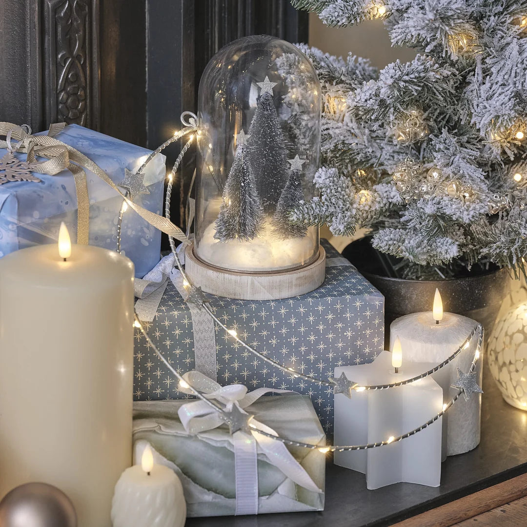 Weihnachts-Schneekugel mit LED-Lichtern, verschneiter Weihnachtsbaum-Szene im Glas-Dome, auf Geschenkbox mit blauem Weihnachtsmuster, warmweiße Lichterkette, aufgeputzte Dekoration mit Schleife.