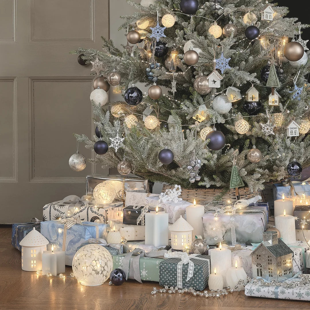 Komplette Weihnachtsdekoration mit geschmücktem Weihnachtsbaum, beige Tannenzweigen, Glaskugeln in Natur- und Blautönen, Schneeflocken-Ornamenten, LED-Kerzen, Porzellan-Häuser mit warmweiß beleuchteten Fenstern, Geschenke, Lichterkette, festliches Wohnzimmer-Styling.