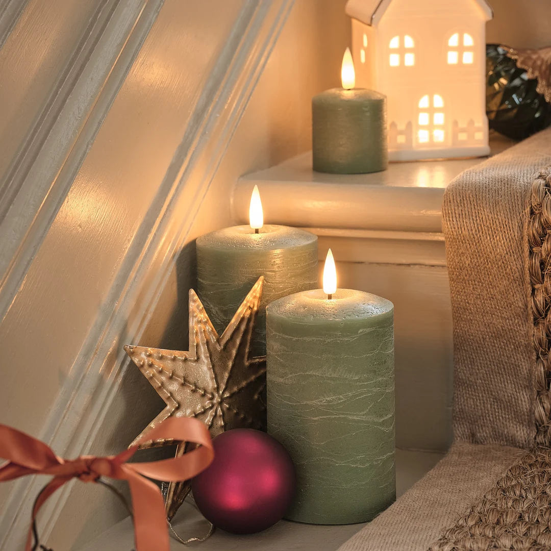Zwei grüne LED-Kerzen mit goldener Stern-Deko und rote Weihnachtskugel, Kerzenhaus mit beleuchteten Fenstern, auf beiger Leinenserviette mit Weihnachtszweigen.