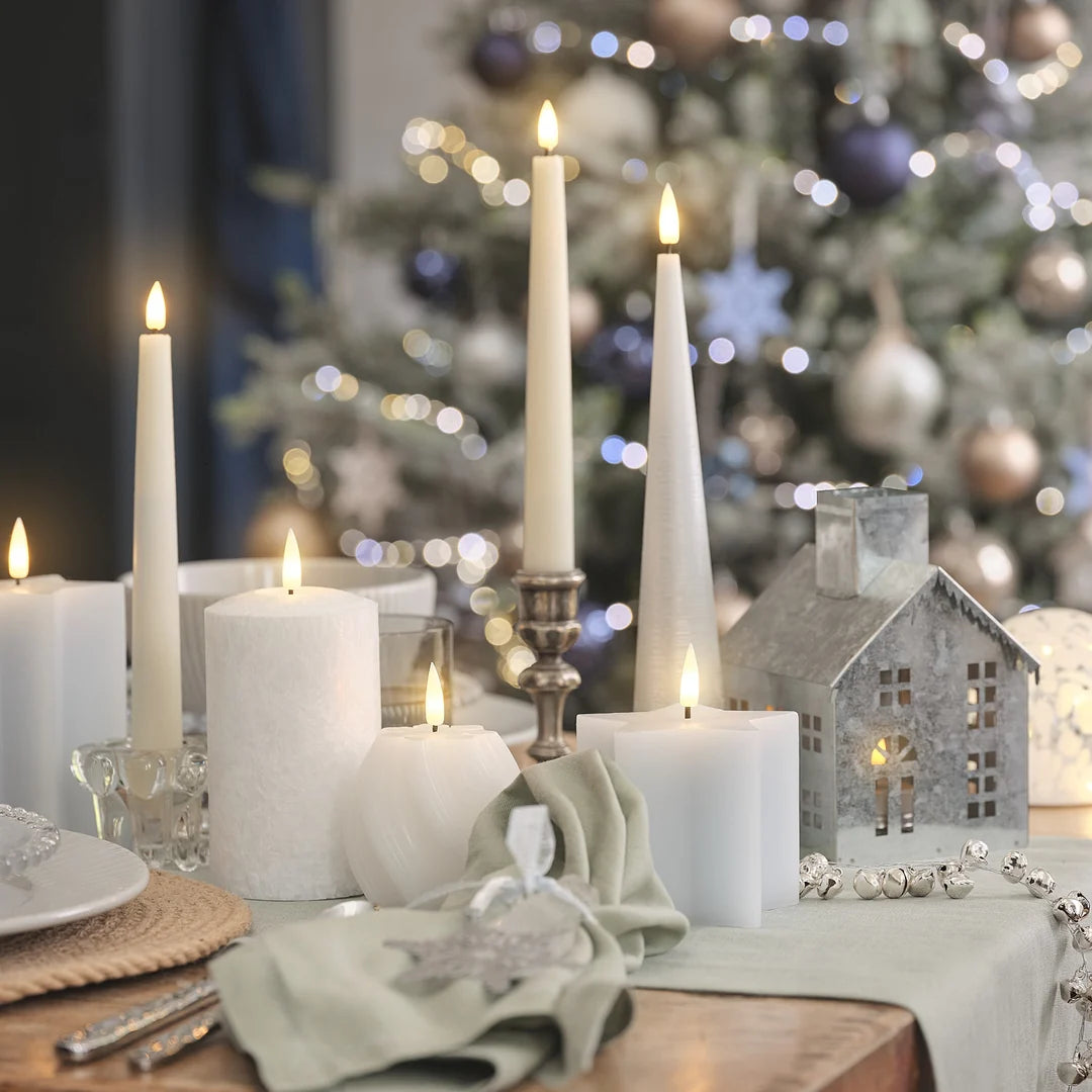 Elegantes Weihnachts-Tischgedeck mit weißen LED-Kerzen, Leuchtern, grünem Leinenservietten, Weihnachtskugeln, Kristall-Gläsern und Dekoration vor unscharfem Weihnachtsbaum.