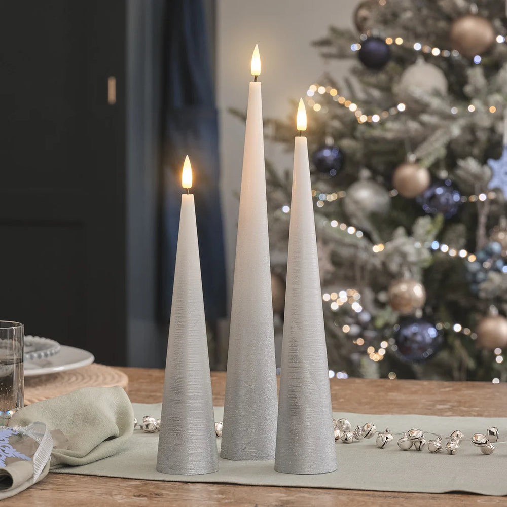 Drei weiße LED-Kerzen in hoher Kegel-Form mit strukturierter Oberfläche und warmweißen LED-Flammen auf Holztisch, beige Leinenserviette, verschwommener Weihnachtsbaum mit roten Kugeln im Hintergrund.