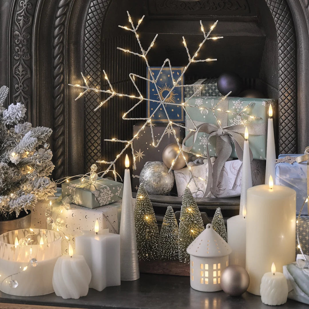 Kaminverzierung mit vier weißen LED-Kerzen in hoher Kegel-Form mit strukturierter Oberfläche, warmweißen LED-Flammen, Weihnachtsgirlande mit grünem Laub, roten Beeren, Zapfen, Weihnachtskugeln in Rot und Gold, weiße Taper-Kerzen auf Kerzenständer, Holzoberfläche.