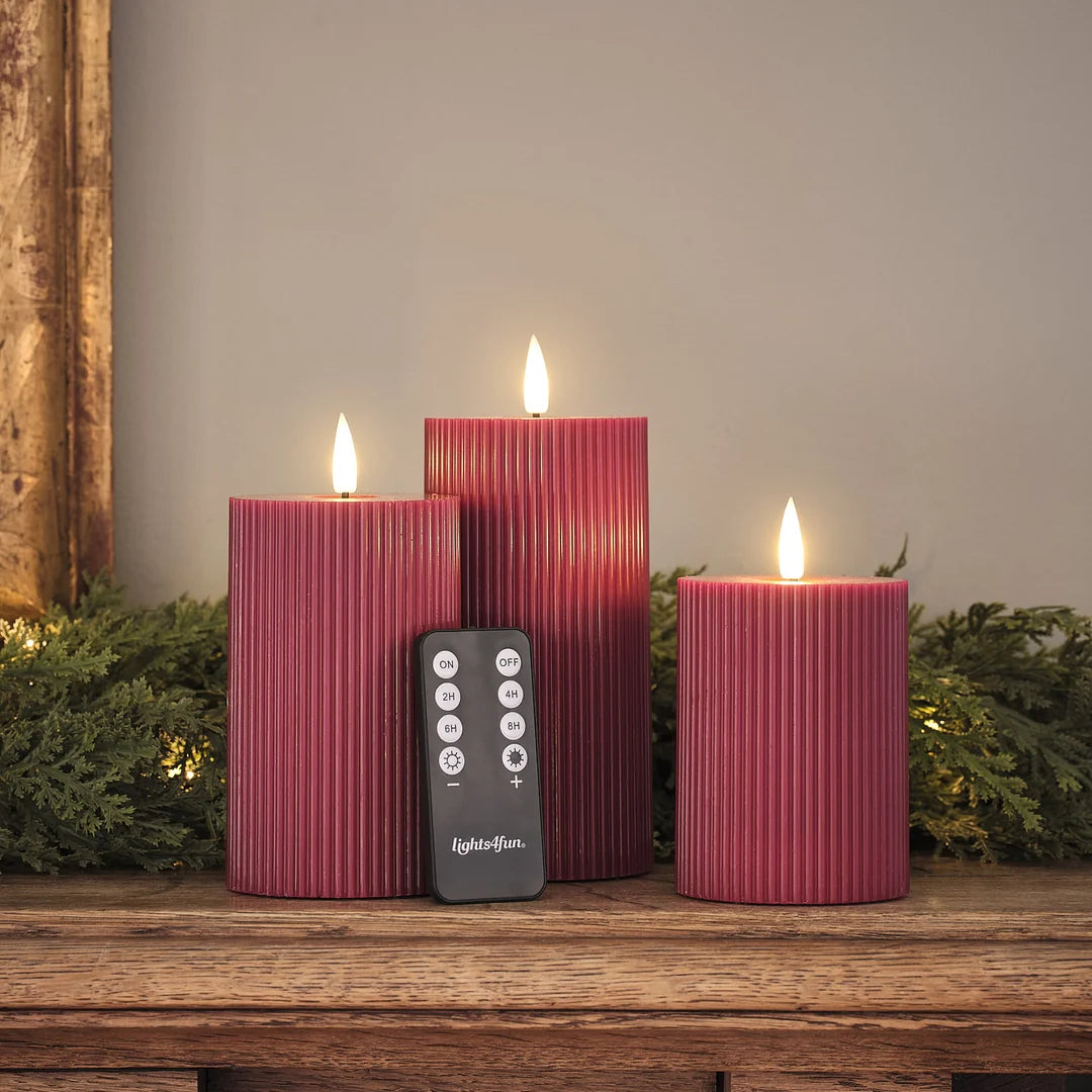 Drei rote LED-Zylinderkerzen mit strukturierter Oberfläche, warmweiße LED-Flammen, schwarze Fernbedienung mit ON/OFF und Timer-Funktionen (2H, 4H, 6H, 8H) auf Holztisch, grüne Girlande.
