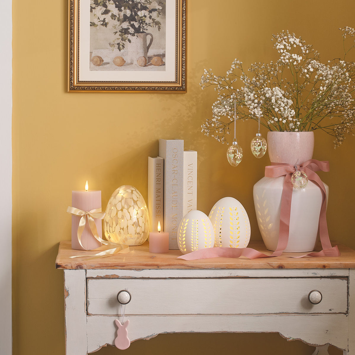 Osterdeko mit Glas-Ostereiern, Kerzen und Büchern auf einem weißen Sideboard vor gelber Wand