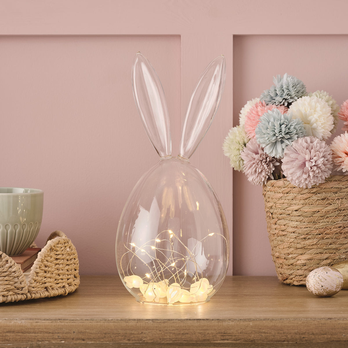 Glaskuppel in Hasenform mit Mikro-LED-Lichterkette auf Holztisch neben Korb mit pastellfarbenen Blumen
