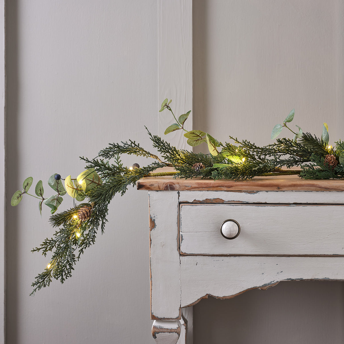 Weihnachtsgirlande mit Tannenzapfen, Beeren und warmweißen Micro-LEDs auf einem weißen Holzsideboard