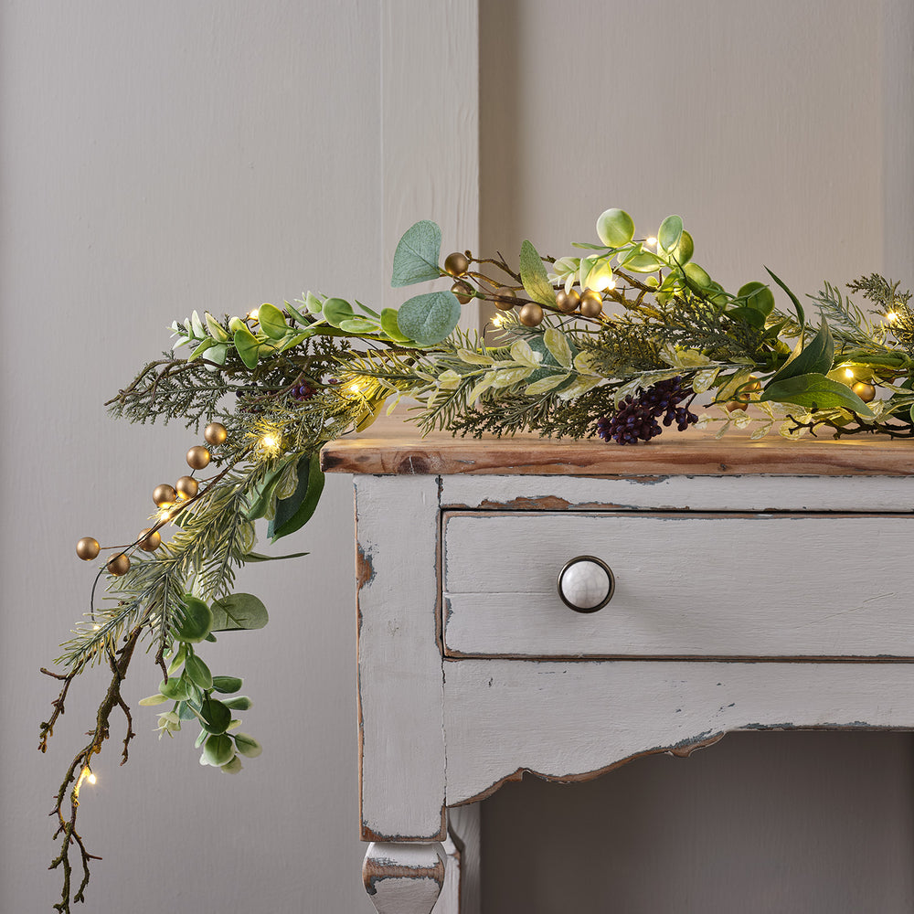 Weihnachtsgirlande mit goldenen Beeren und grünen Blättern dekorativ auf einem weißen Holzsideboard mit Micro-LED-Lichtern