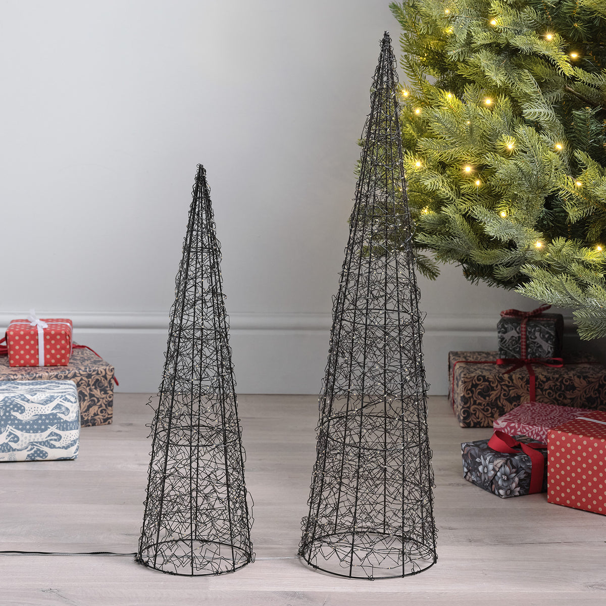 Unbeleuchtete LED-Kegel aus Metalldraht in zwei Größen auf Holzboden neben Weihnachtsbaum und Geschenken