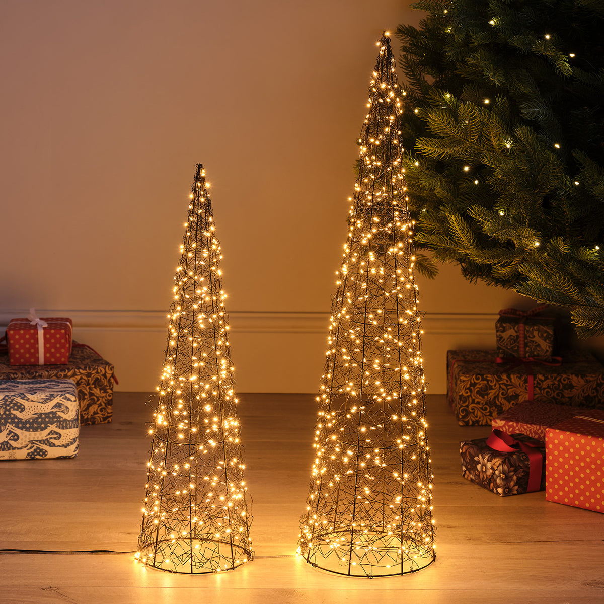 Zwei beleuchtete LED-Kegel aus Metalldraht in verschiedenen Größen vor Wand mit Weihnachtsbaum und Geschenken