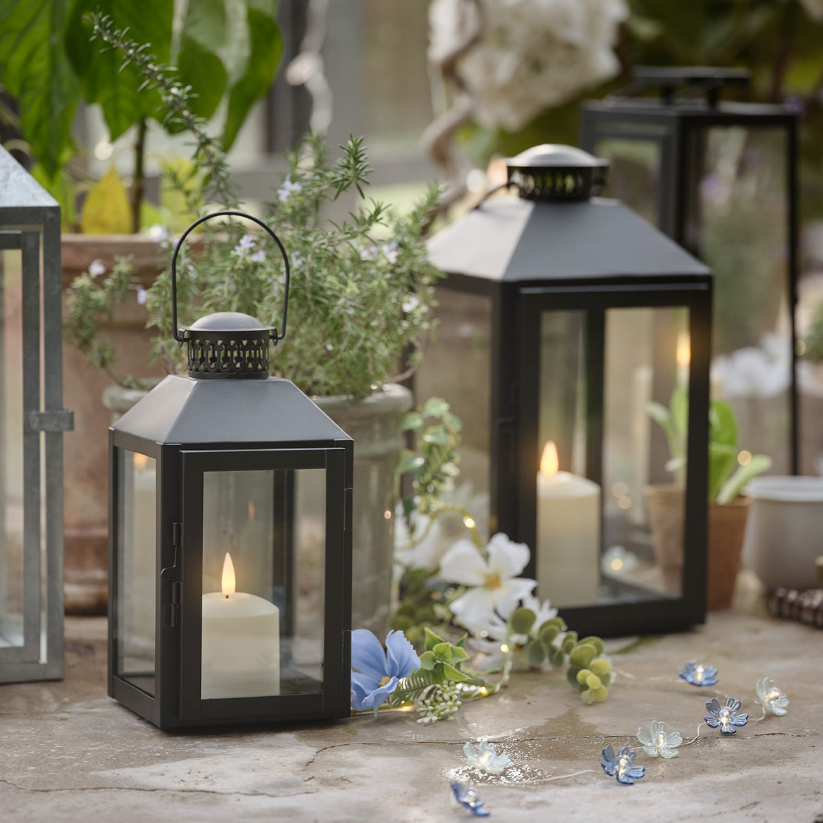 Zwei schwarze Laternen mit TruGlow LED-Kerzen und Blumendekoration auf Steinboden