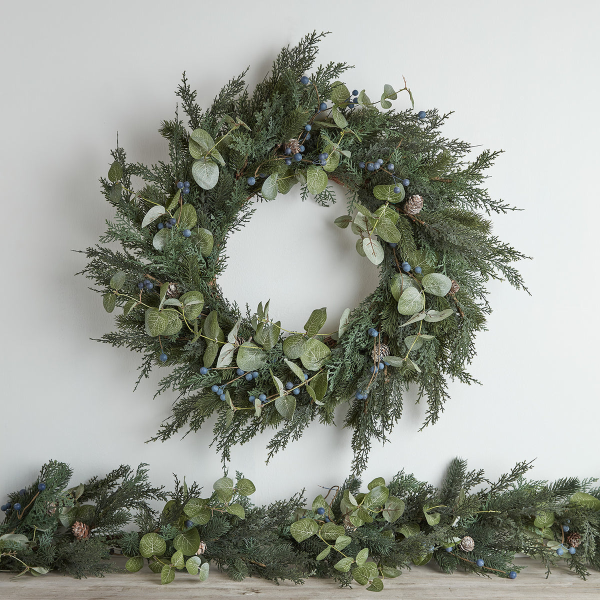 Weihnachtskranz ohne Beleuchtung mit grünen Tannenzweigen, Eukalyptusblättern, blauen Beeren und Tannenzapfen an weißer Wand